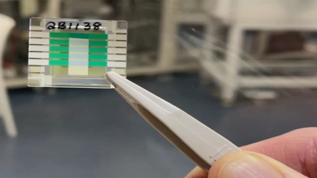 A hand holds tweezers with a small solar cell sample in a lab setting.