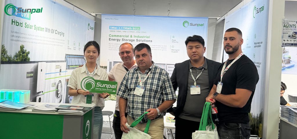 A group photo of five people inside the Sunpal booth at The Smarter E Europe 2026. Two Sunpal representatives stand alongside three European visitors who are holding green Sunpal tote bags, with commercial and industrial energy storage solution banners visible in the background.